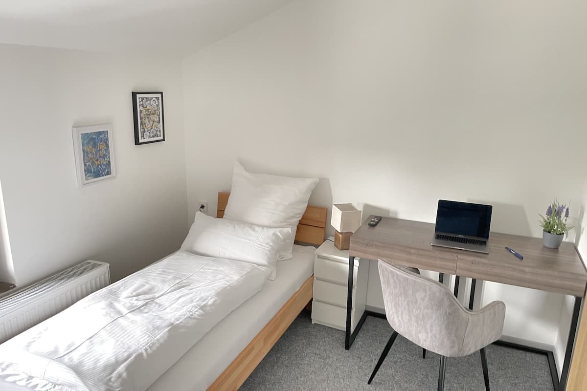 Quarto bem arrumado com uma cama, uma escrivaninha, uma cadeira, um laptop e uma pequena planta em vaso.