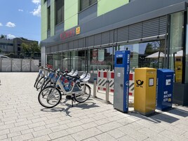 Un edificio con un letrero que dice "ECKERT" y un anuncio de lotería. Hay bicicletas estacionadas al frente, y una caja azul y amarilla con texto.