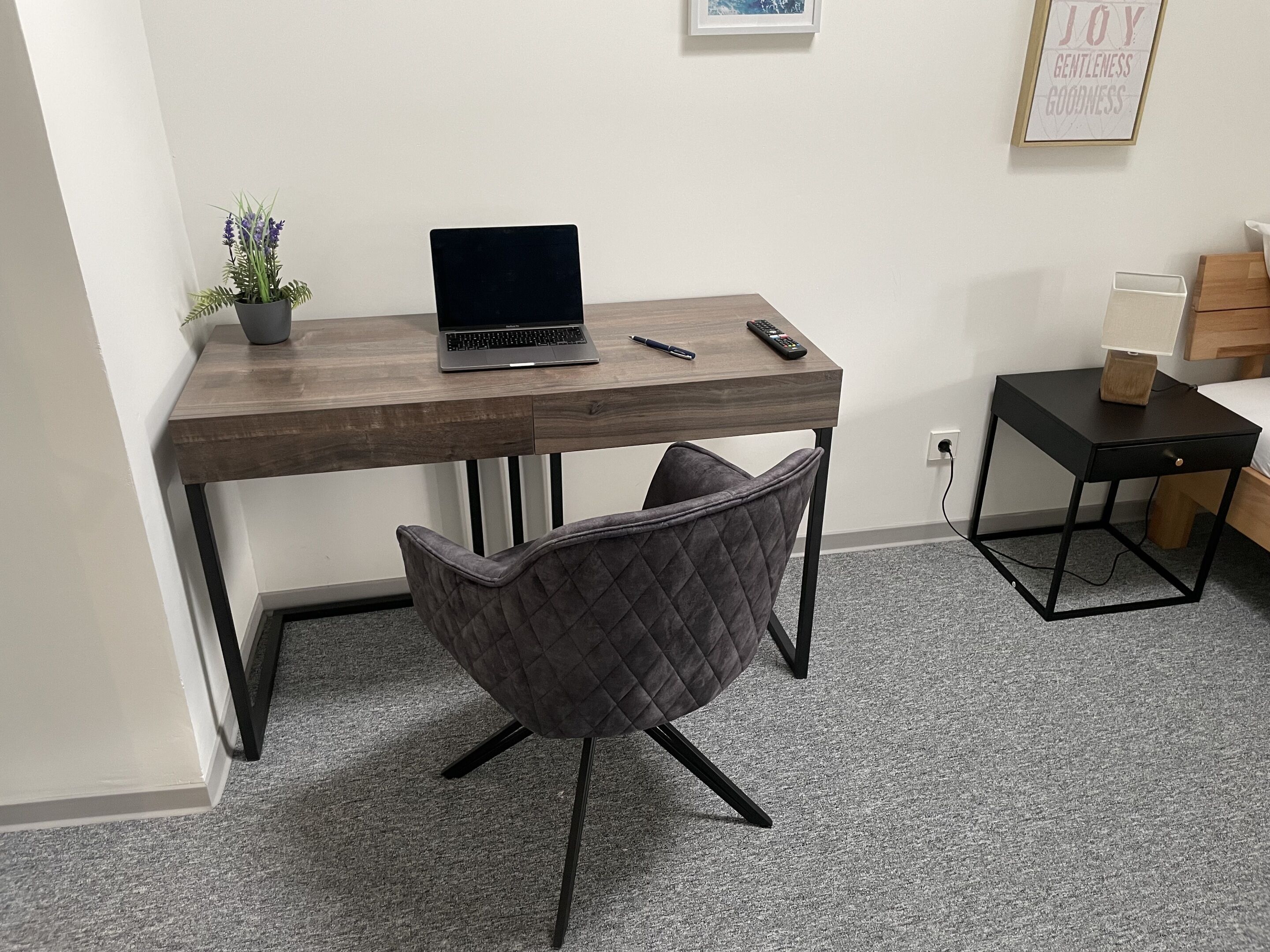 Ein kompakter Arbeitsbereich mit einem Laptop auf einem Holztisch, einem grauen Stuhl und einem kleinen Tisch mit Kartons.