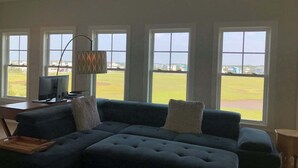 Una sala de estar con un sofá modular, un ventilador de techo y ventanas amplias con vistas a un campo verde.