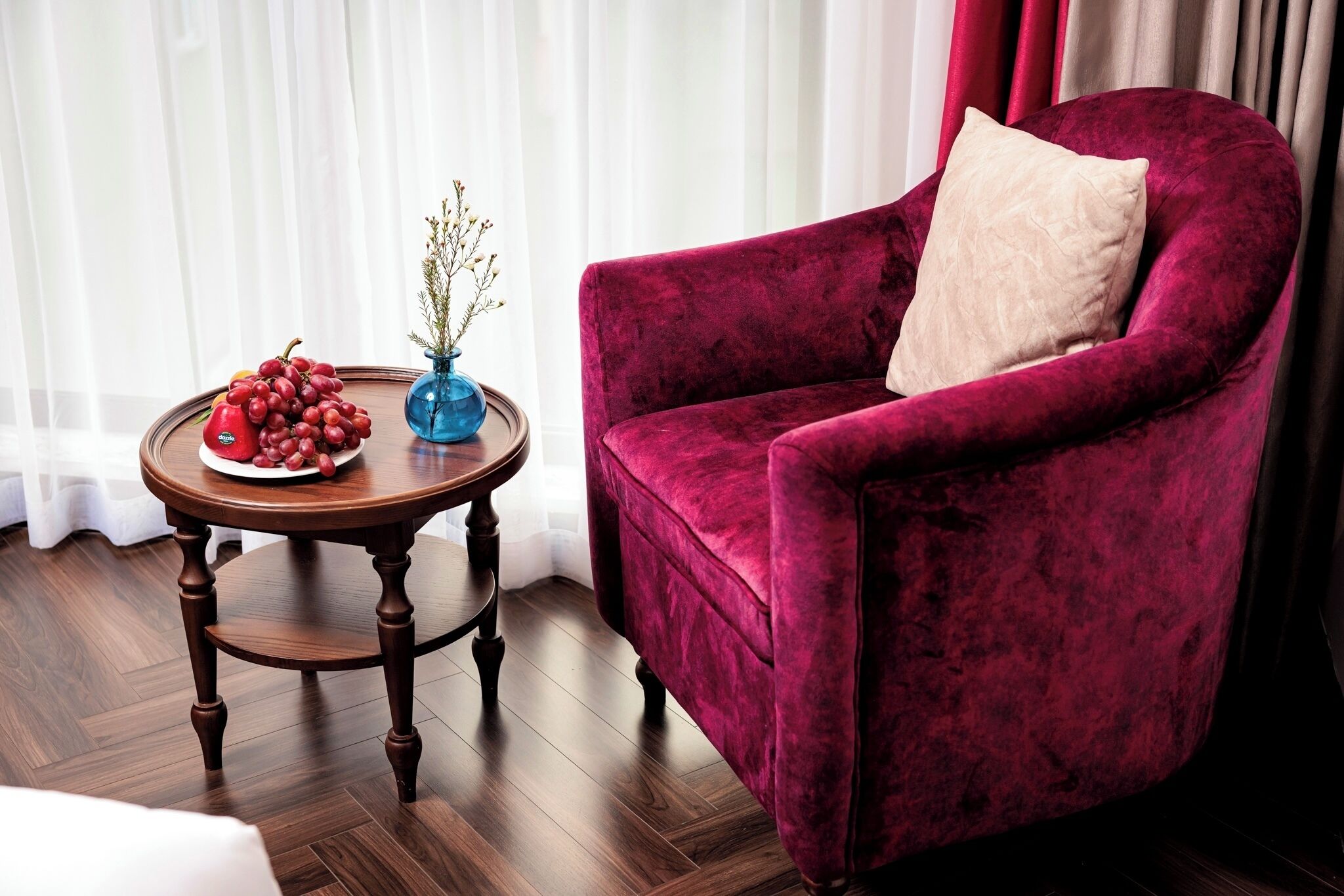 A plush red velvet armchair with a beige pillow, a wooden side table with a blue vase and fruit, and a wooden floor.