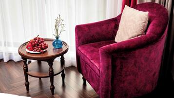A plush red velvet armchair with a beige pillow, a wooden side table with a blue vase and fruit, and a wooden floor.