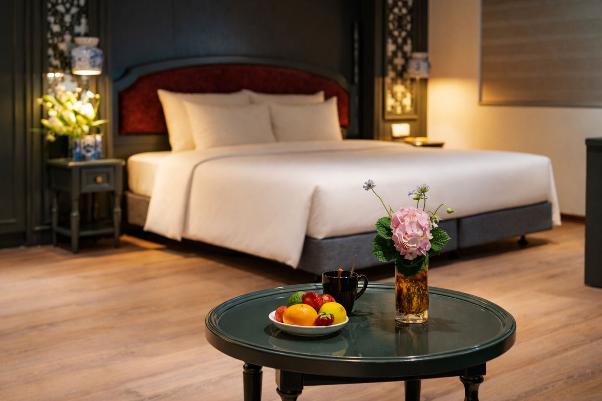 A hotel room with a bed, a round table with fruit and a drink, and a vase with flowers.