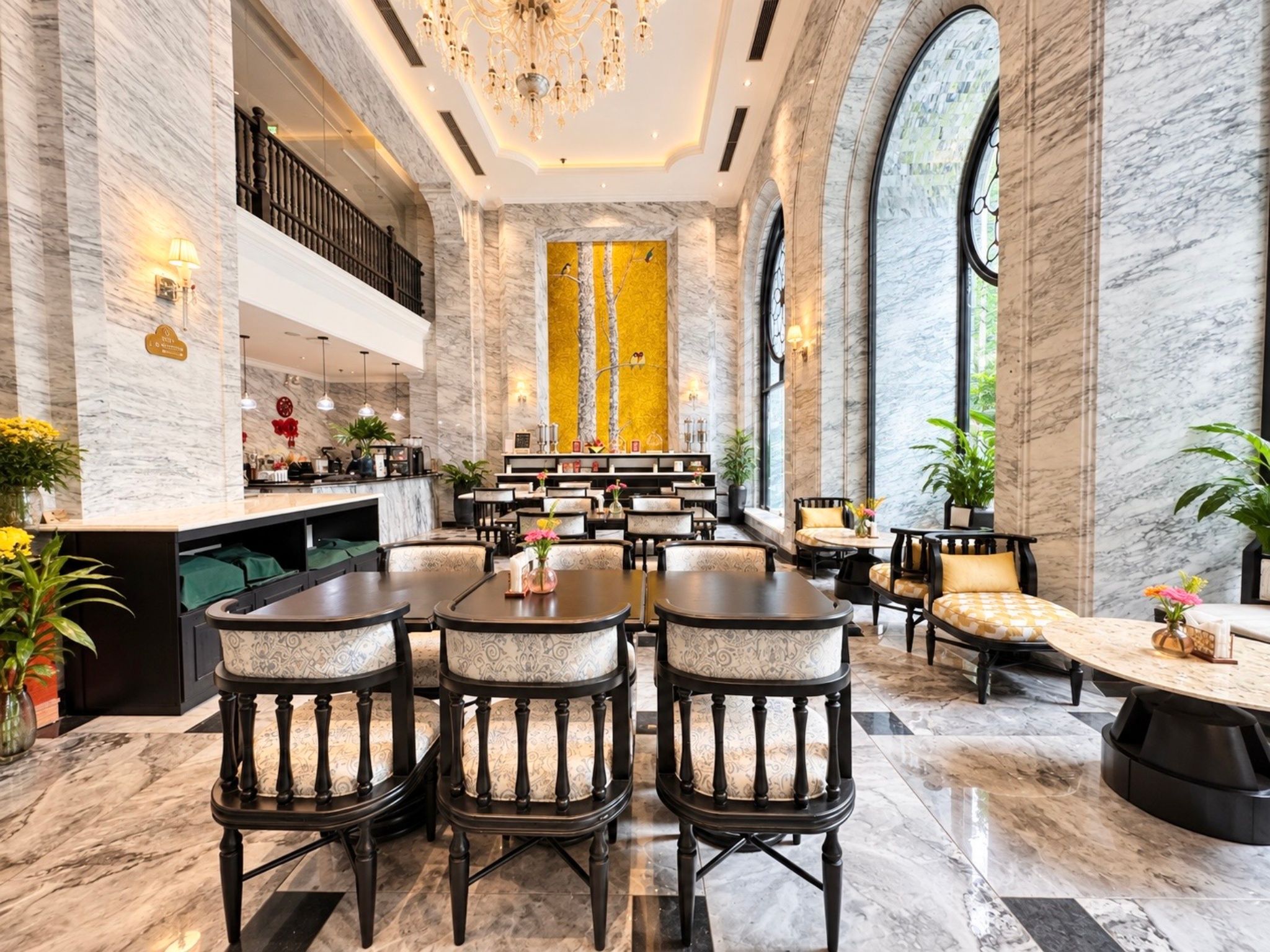 A spacious hotel lobby with marble floors, a central seating area with tables and chairs, a reception desk, and a large arched window.