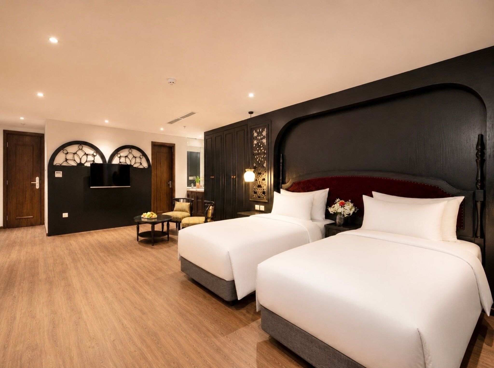 Ein Hotelzimmer mit zwei Betten, einer Sitzecke mit Tisch und Stühlen, einem an der Wand montierten Fernseher und einem dunklen Kopfteil.
