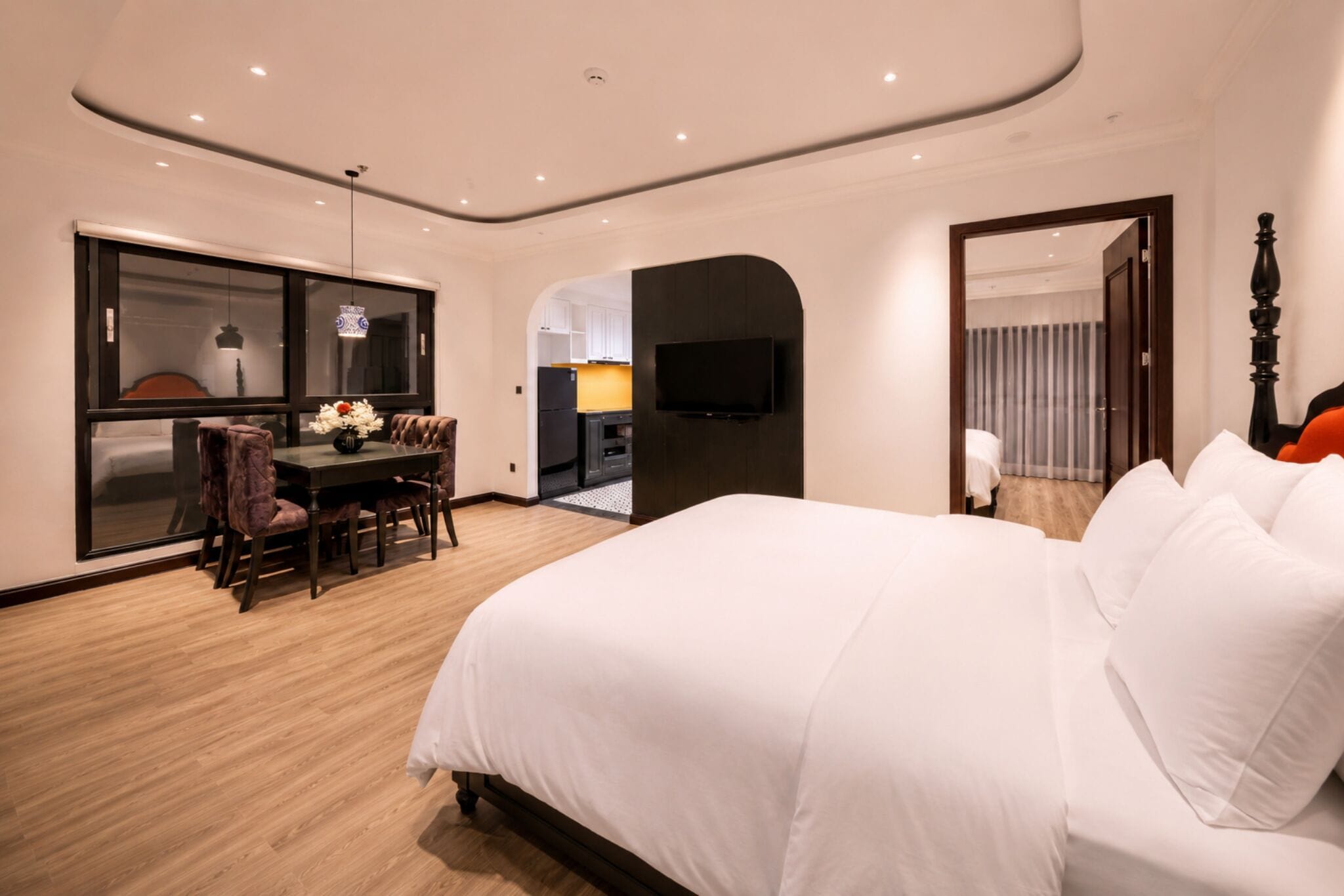 A modern hotel room with a large bed, a dining area, a TV, and a view of the kitchen.