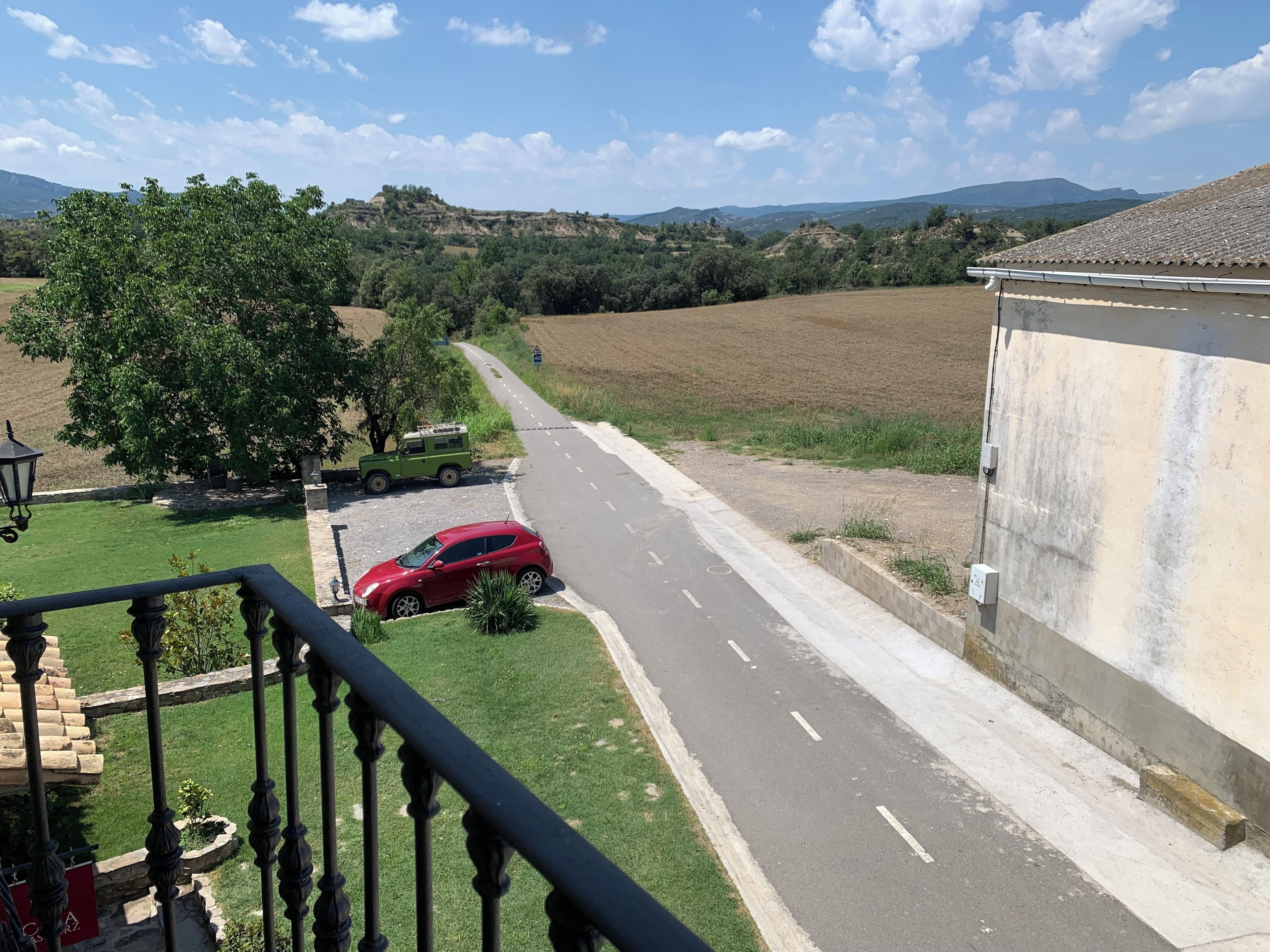 Una strada con un'auto rossa parcheggiata a lato, un veicolo verde e un edificio con tetto in tegole.