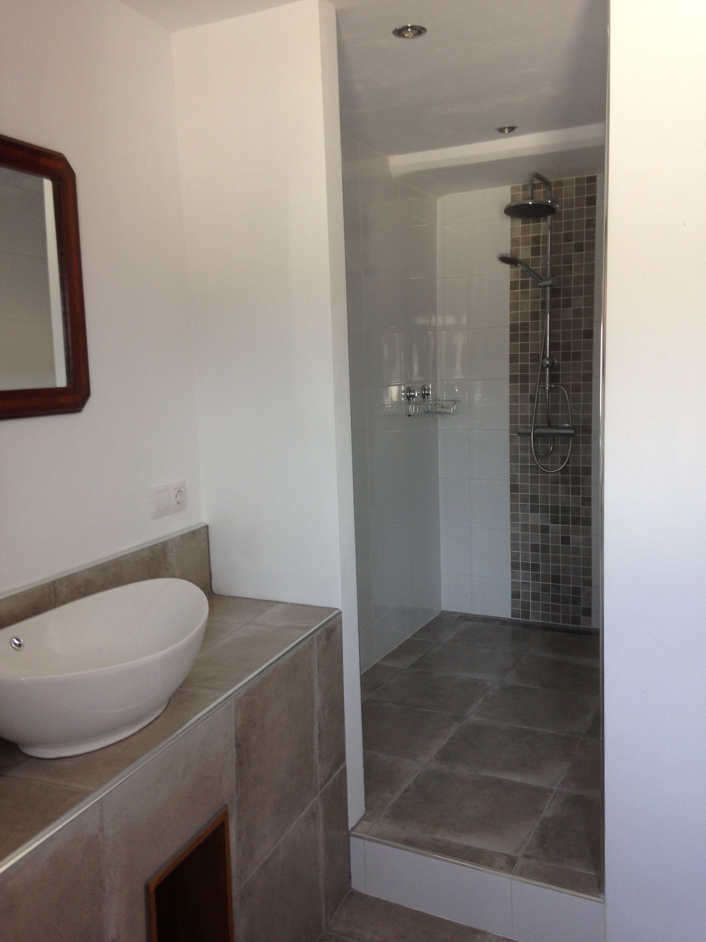 Une salle de bain avec un grand lavabo blanc, un miroir et une douche dont les murs et le sol sont recouverts de carreaux.