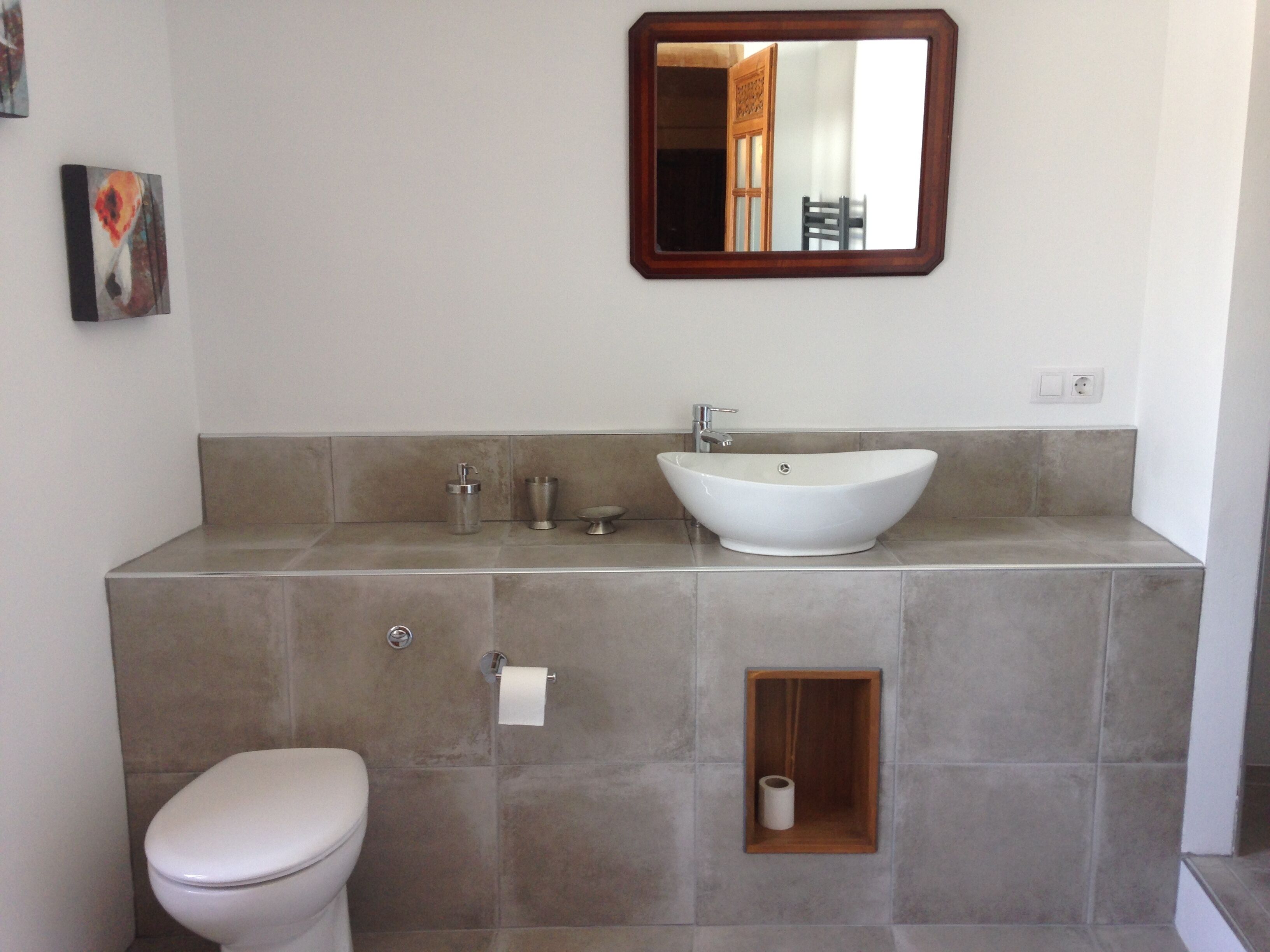 Un baño con inodoro blanco, lavabo sobre encimera, espejo y pequeño estante de madera con dispensador de papel higiénico.