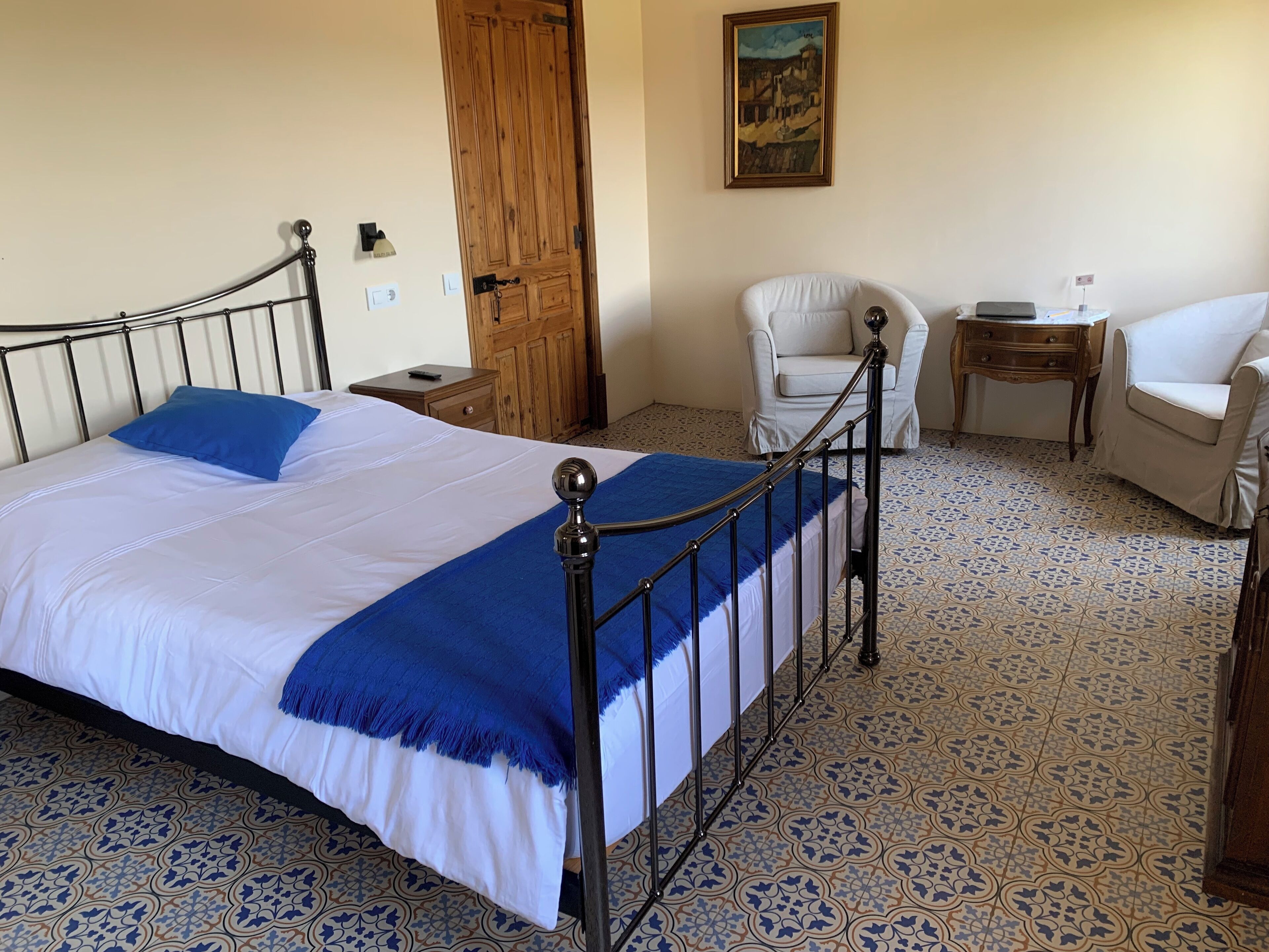 Una cama bien hecha con colcha azul y blanca, una puerta de madera, un sillón blanco, una mesita de noche de madera y un suelo con diseño.
