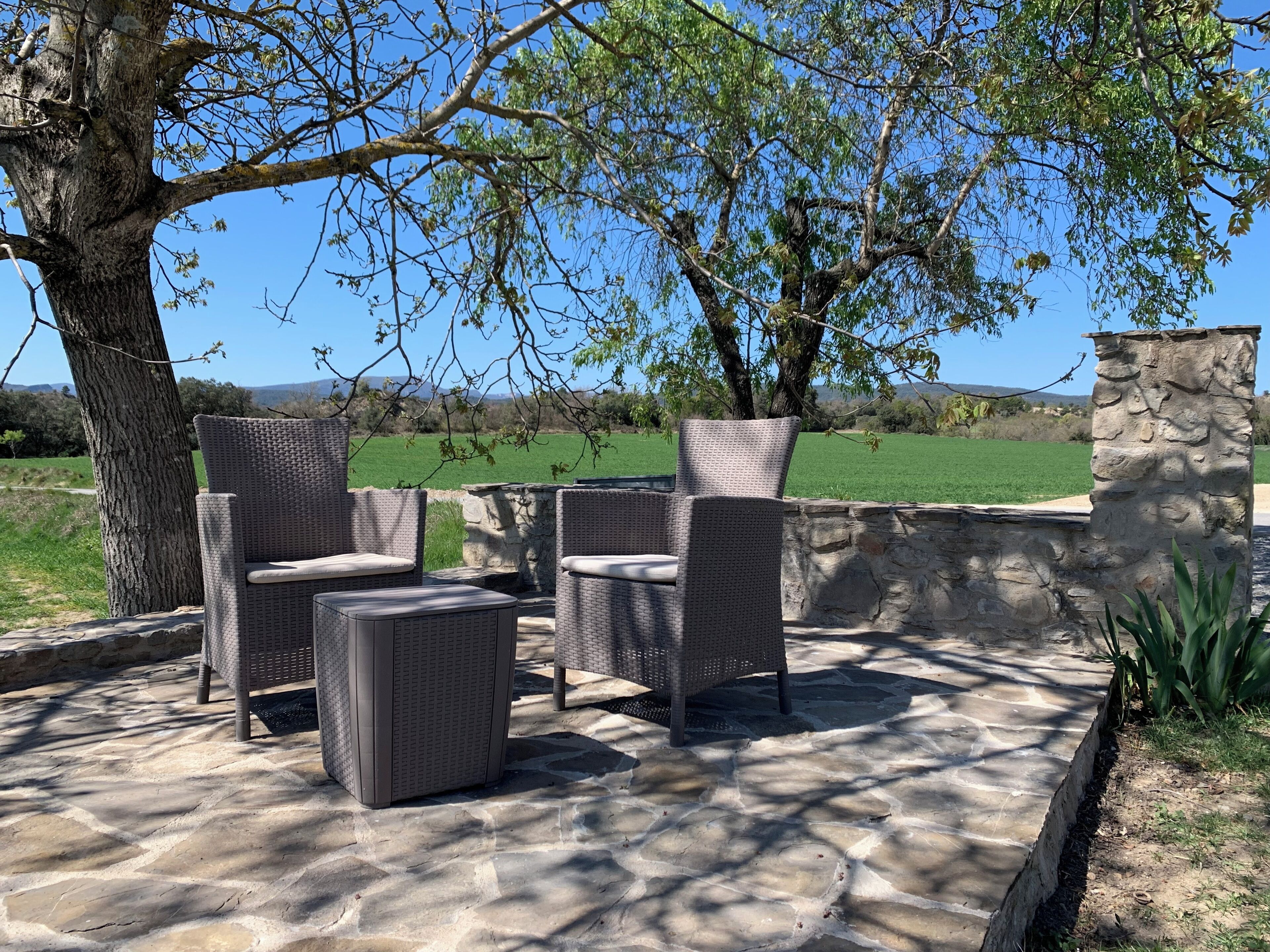 Een terras buiten met rieten meubels, stenen vloeren en uitzicht op een groen veld.