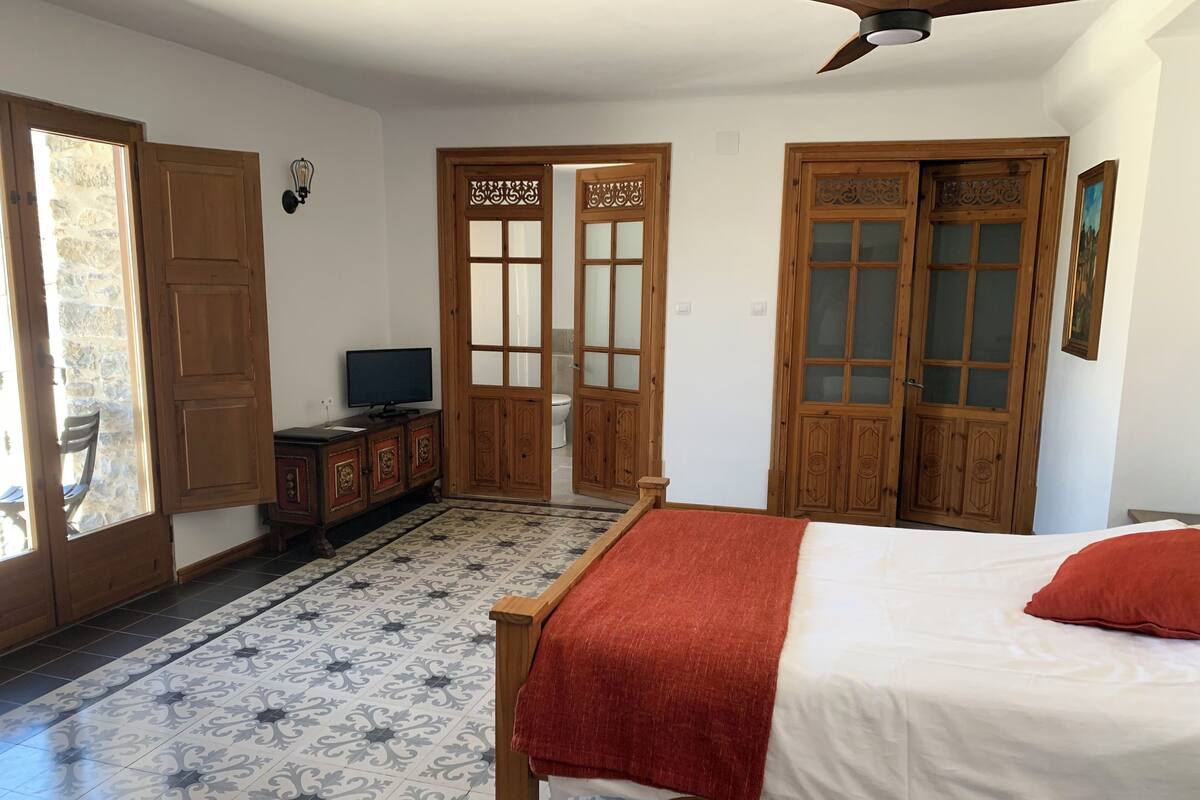 A bedroom with a bed, a television on a wooden cabinet, and a bathroom visible through a wooden door.