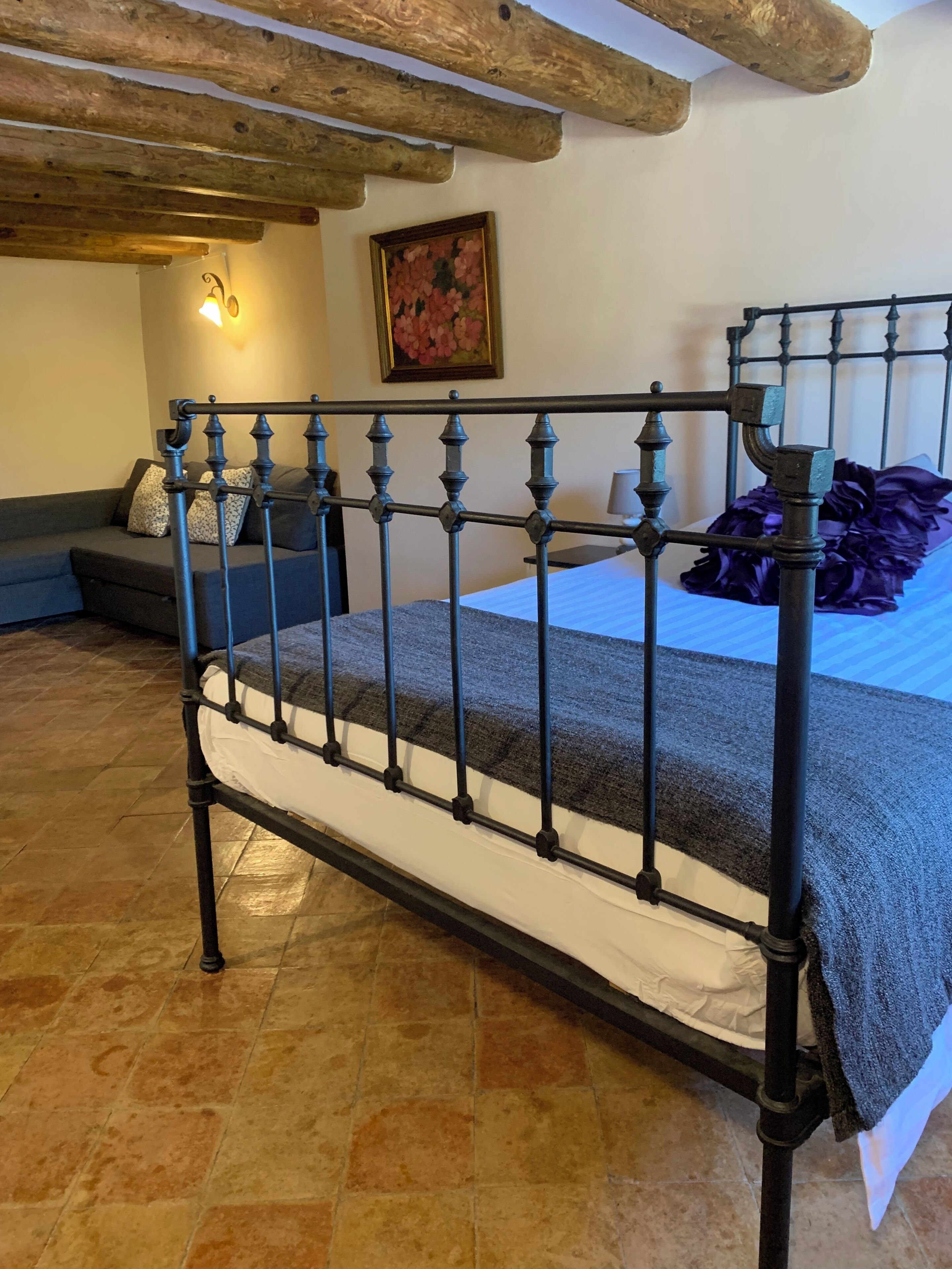 Una cama con estructura metálica, una manta gris y un cojín morado.