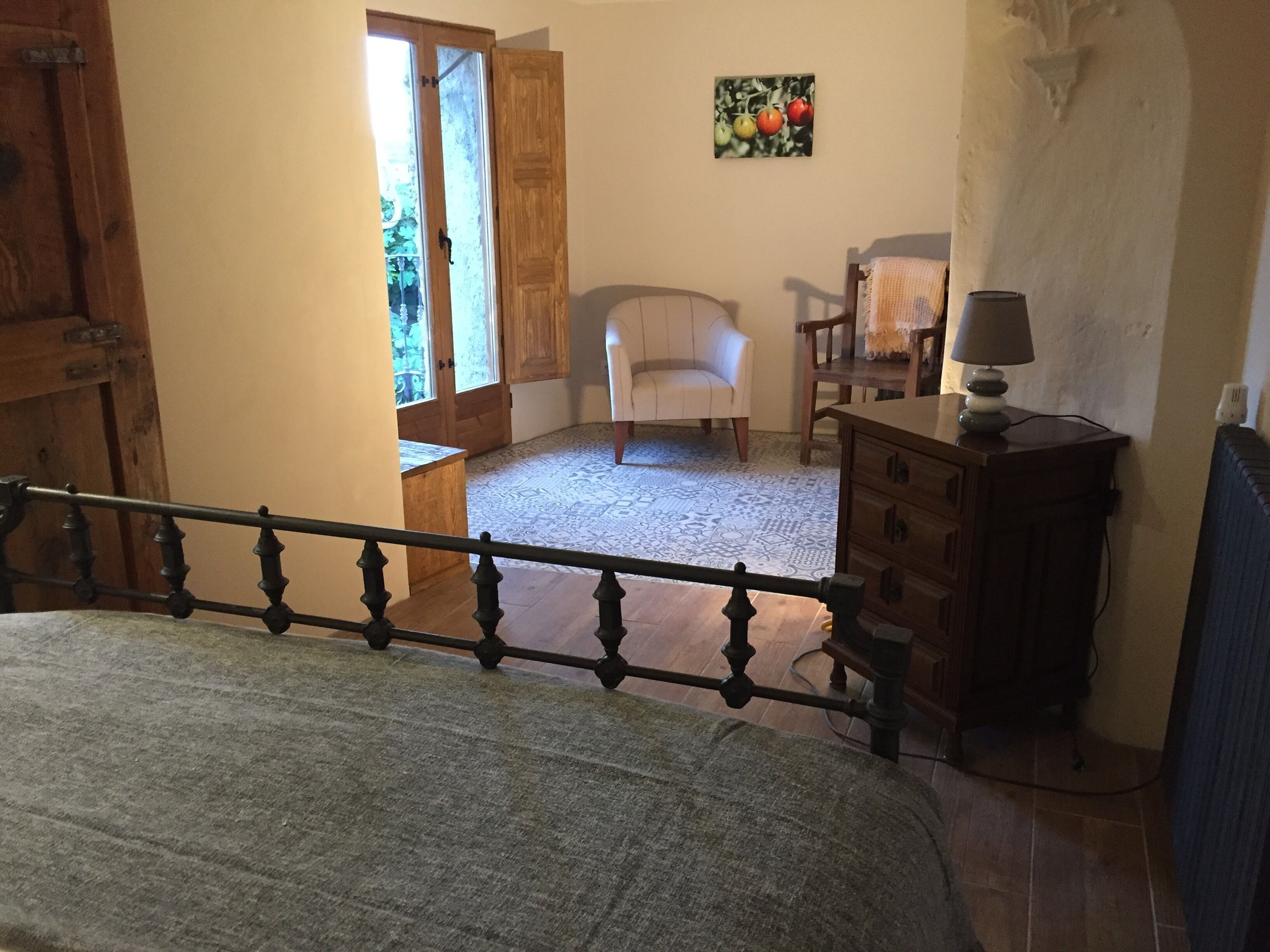 Un dormitorio con cama, cómoda, silla y mesa. Se ve follaje verde por la ventana.