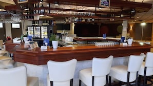 A bar area with a wooden counter, multiple glasses, and a potted plant. There are bar stools and a refrigerator in the background. - De Schildkamp (Asperen)