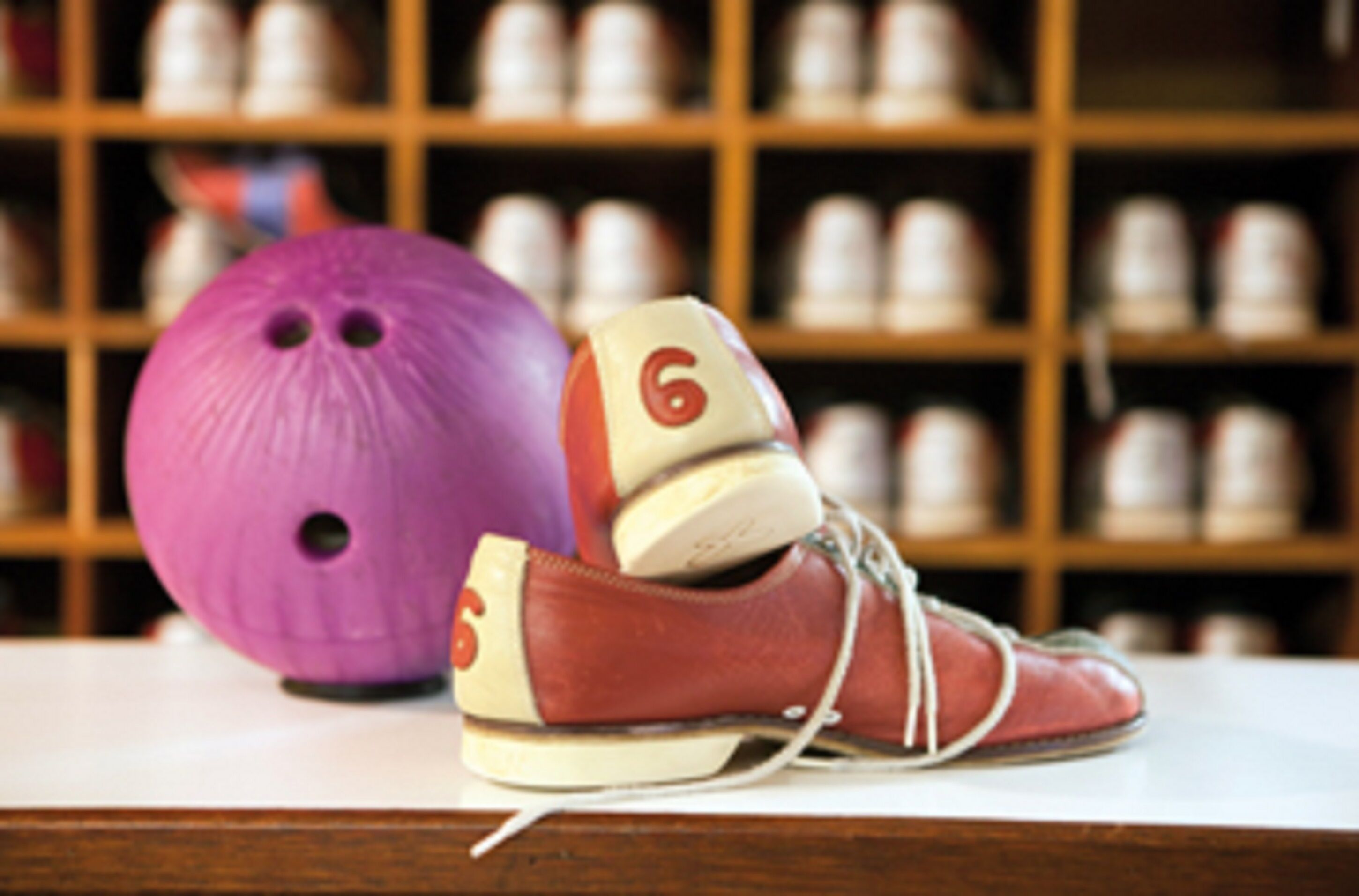 Una bola de bitlles morada i un parell de sabates de bitlles vermelles amb el número 6, damunt una superfície de fusta i un fons desenfocat d'estanteries.