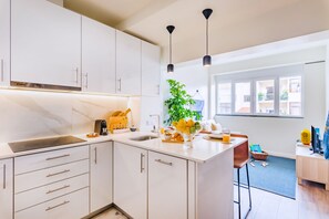 A modern kitchen with white cabinets, a built-in oven, and a large island with a sink. There is a window providing natural light and a view of the outside. - Cascais Modern & Quiet Retreat (Cascais)