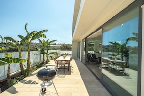 A modern patio with a grill, dining table, and chairs, surrounded by glass walls and tropical plants.