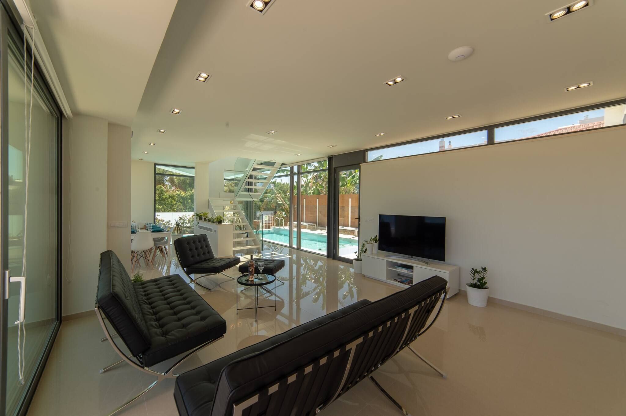 Una sala de estar moderna con televisor de pantalla plana, sofás de cuero negro, una mesa de comedor de vidrio y vista a la piscina.