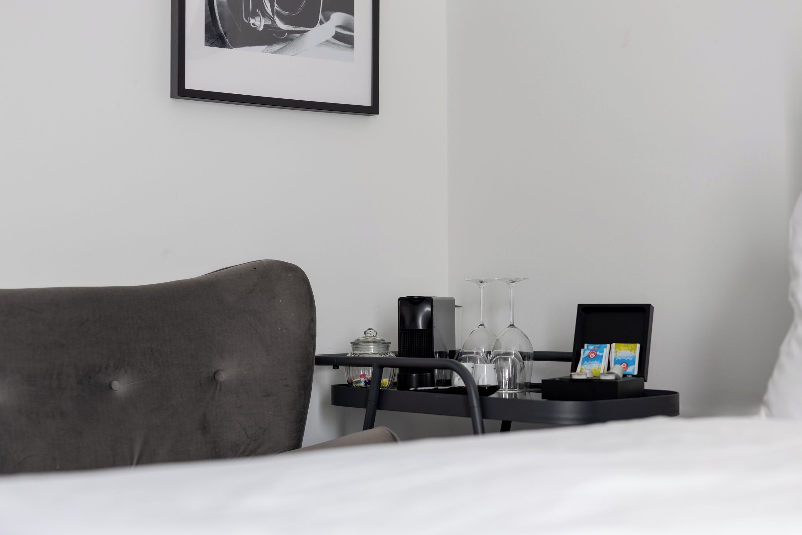 Habitación de hotel ordenada con una cama, una mesita de noche con un cuadro enmarcado, una pequeña bandeja negra con vasos y una tarjeta, y una lámpara de noche.