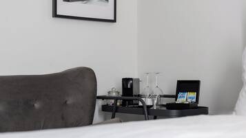 Une chambre d’hôtel bien rangée, avec un lit, une table de chevet sur laquelle est posée une photo encadrée, un petit plateau noir avec des verres et une carte, et une lampe de chevet.