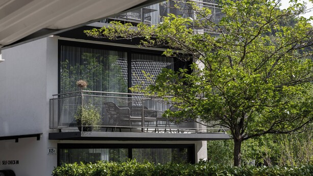 Un edificio moderno con balcón, zona de registro automático y abundante vegetación.