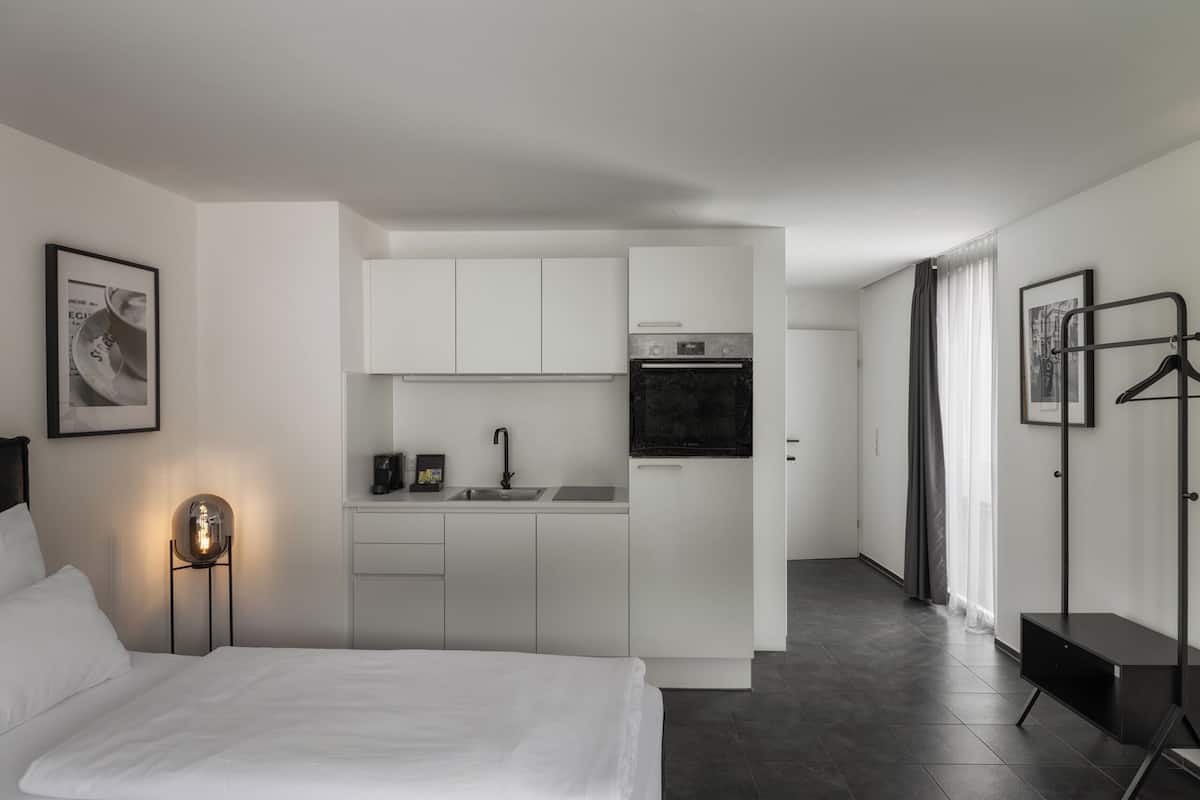 A compact hotel room with a bed, a small kitchenette, a bedside table with a lamp, and a wardrobe.