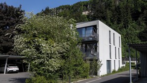 Un edificio moderno con un porche cubierto, rodeado de abundante vegetación y una ladera.