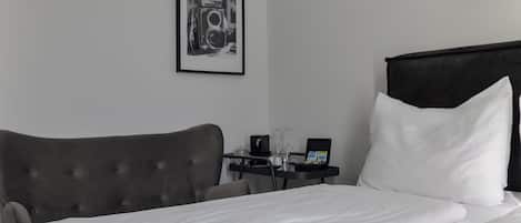 Una habitación de hotel con una cama bien hecha, un sillón tapizado en gris, una mesita de noche con un vaso, un cuadro enmarcado y un cartel en la pared que dice 'BED NOW'.