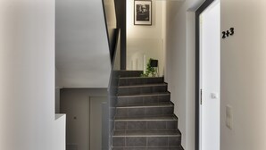 Una escalera moderna con baldosas oscuras, un cuadro enmarcado arriba y una puerta con el símbolo de número.