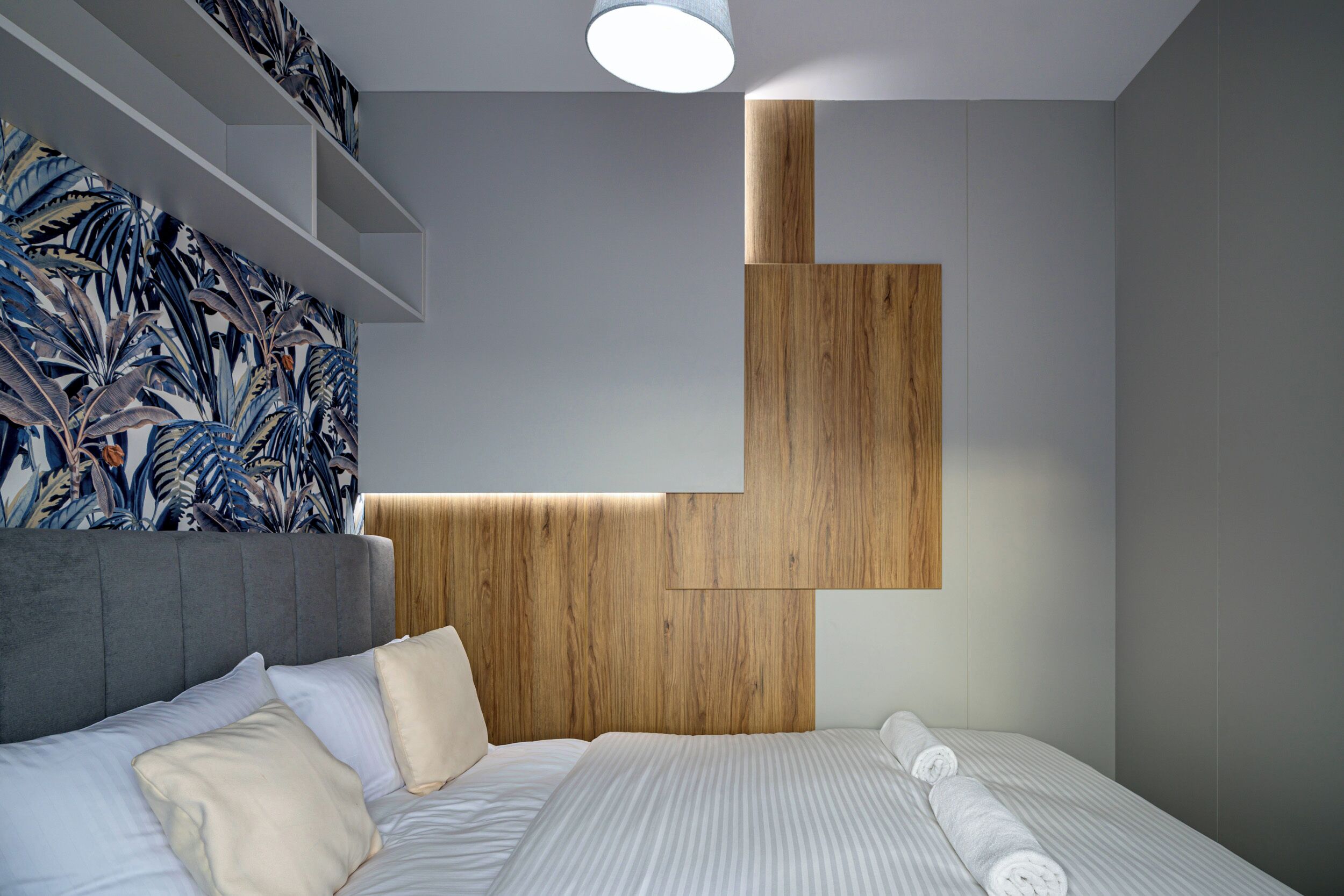 Ein modernes Schlafzimmer mit grauem Stoffpolster-Kopfteil, einer Wandpaneele aus Holz und einem Bett mit weißen Leinen und aufgerollten Handtüchern.