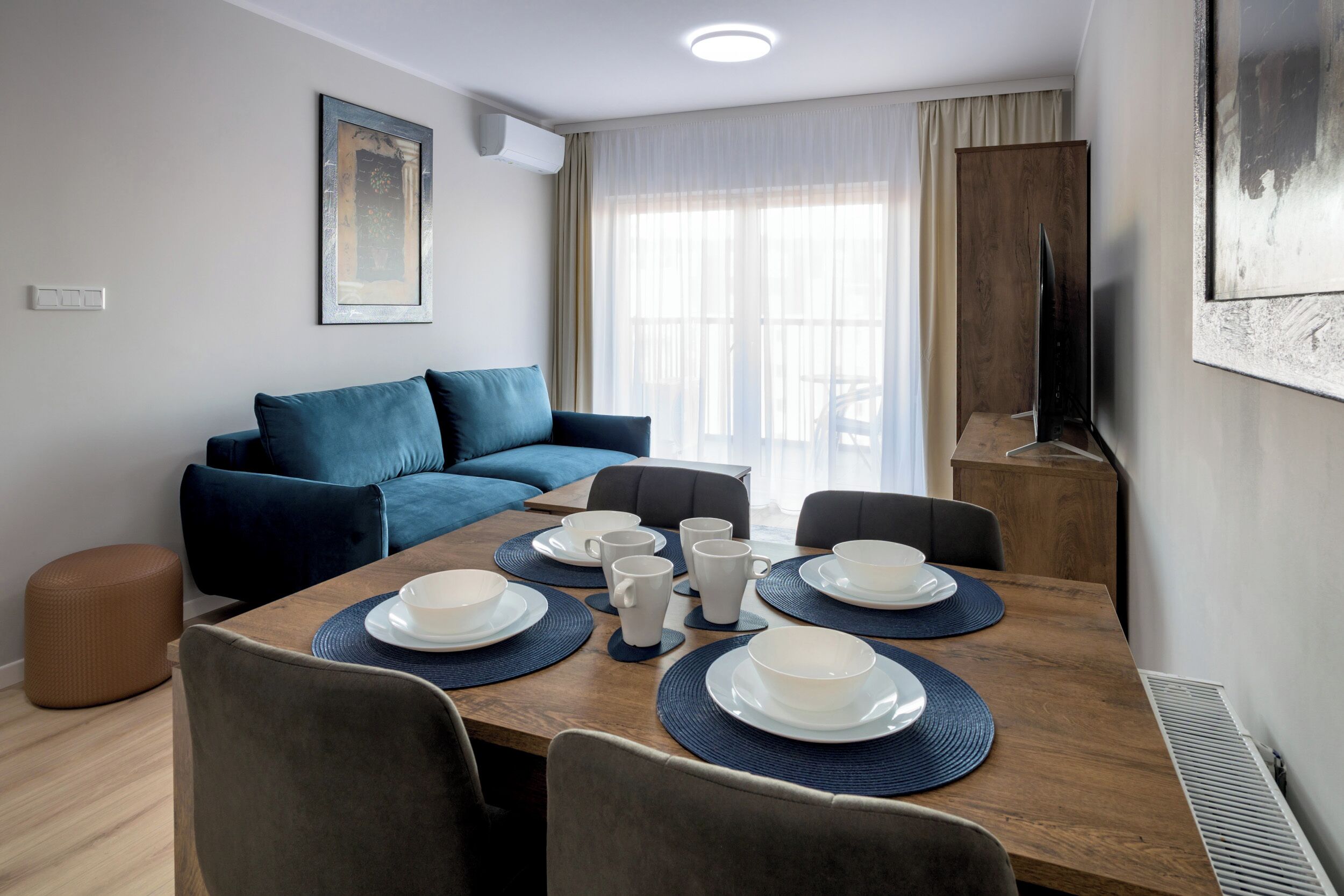 Un salon moderne avec une table à manger dressée pour six personnes, un canapé bleu et un téléviseur posé sur un meuble en bois.
