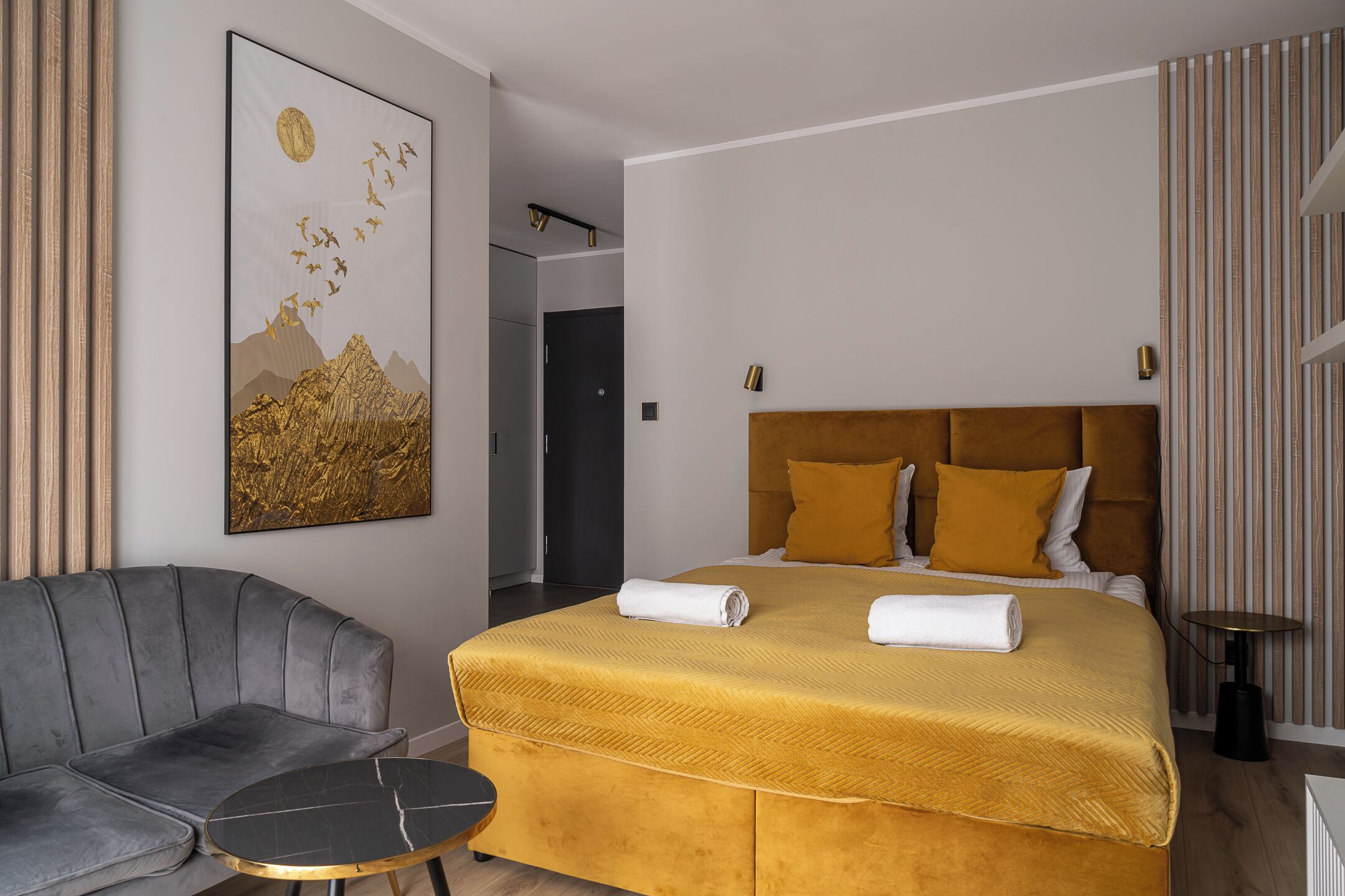 A modern hotel room with a large bed, a gray sofa, a small round table, and a painting on the wall.