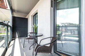 A balcony with two wicker chairs and a glass table, a potted plant, and a view of a building and greenery. - RentPlanet - Atal City Square (Wroclaw)