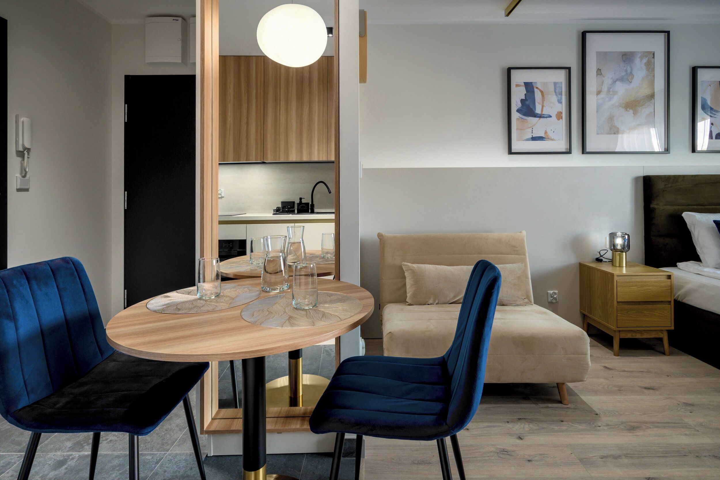 Ein modernes Hotelzimmer mit einem runden Holztisch, zwei blau gepolsterten Sesseln, einem beigen Sofa, einem Nachttisch aus Holz und einem Bett mit Kopfteil.