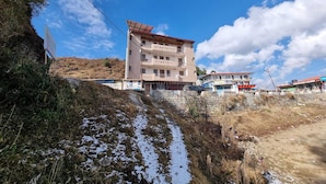 Un edificio de varios pisos con balcones, un sendero de tierra con charcos y un cielo azul despejado.