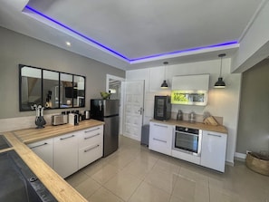 Cozinha moderna com armários brancos, uma geladeira preta, forno embutido e micro-ondas. Possui bancada de madeira, uma pia e um espelho acima dela.