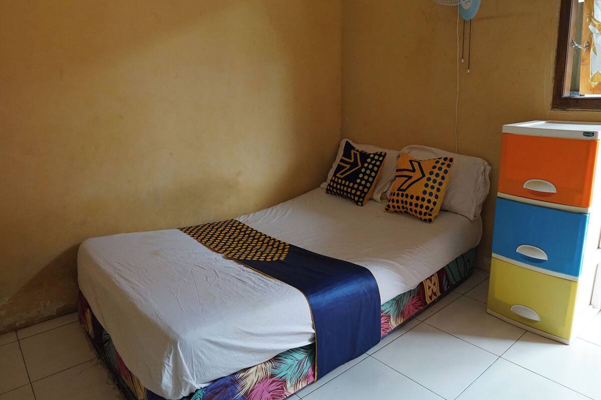 Ein Einzelbett mit weißblauer Bettdecke, zwei gemusterte Kissen und einem farbenfrohen floralen Topper. Ein kleiner Ventilator ist an der Wand über dem Bett angebracht.