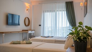 A hotel room with two beds, a TV, a desk, and a window with sheer curtains. - Maslin Otel (Gökçeada)