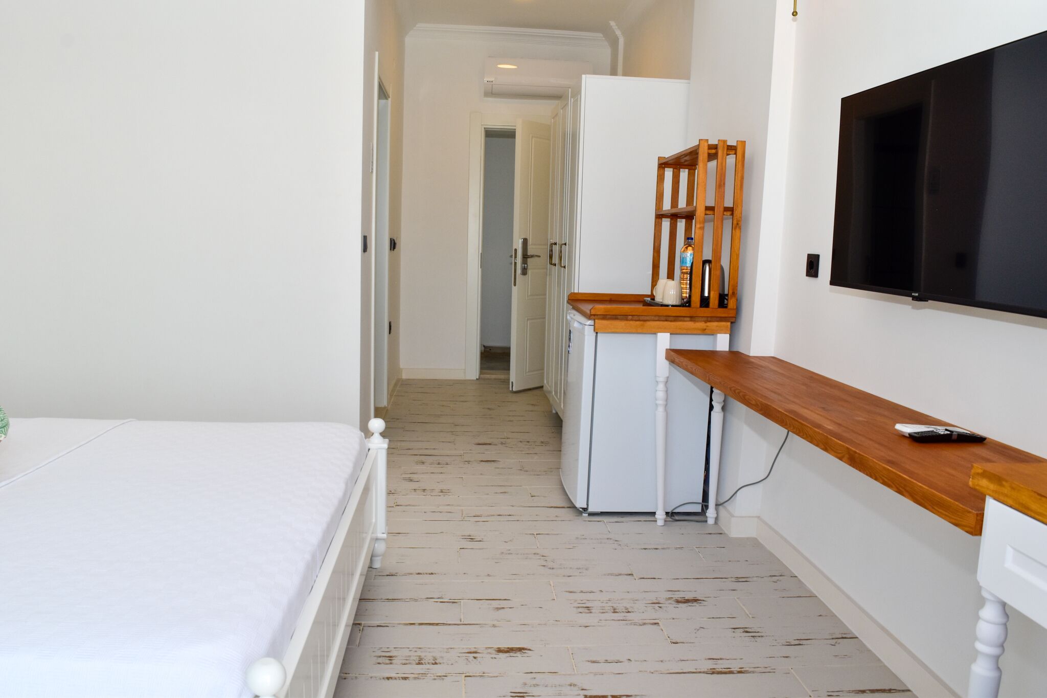 A narrow hallway with a bed on the left, a small kitchenette with a wooden counter and a TV mounted on the wall, and a door leading to another room.