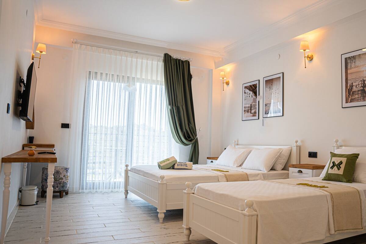 A hotel room with two beds, a wooden floor, a window with sheer curtains, wall-mounted lamps, and framed pictures.