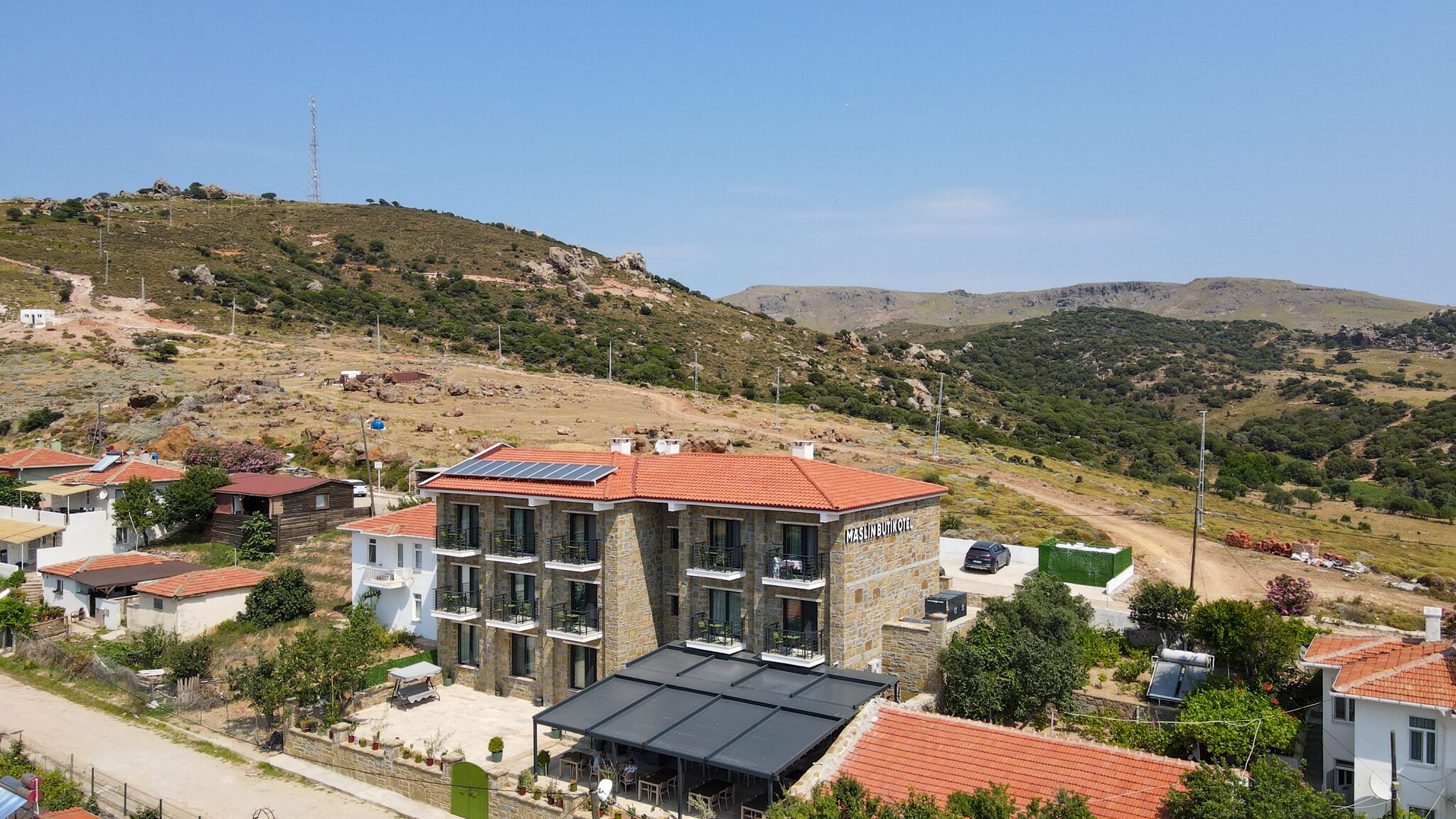 A hillside building with multiple balconies and solar panels, surrounded by residential houses and a hilly landscape.