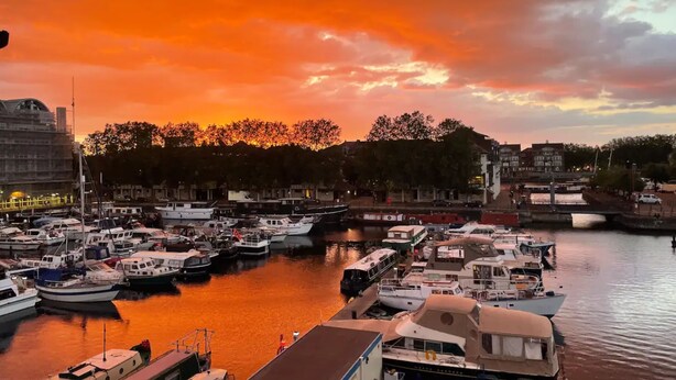 Satamassa on useita laivoja, jotka on ankkuroitu laitureihin. Satamaa ympäröivät rakennukset ja puut, ja taivaalla näkyy auringonlaskun värit.