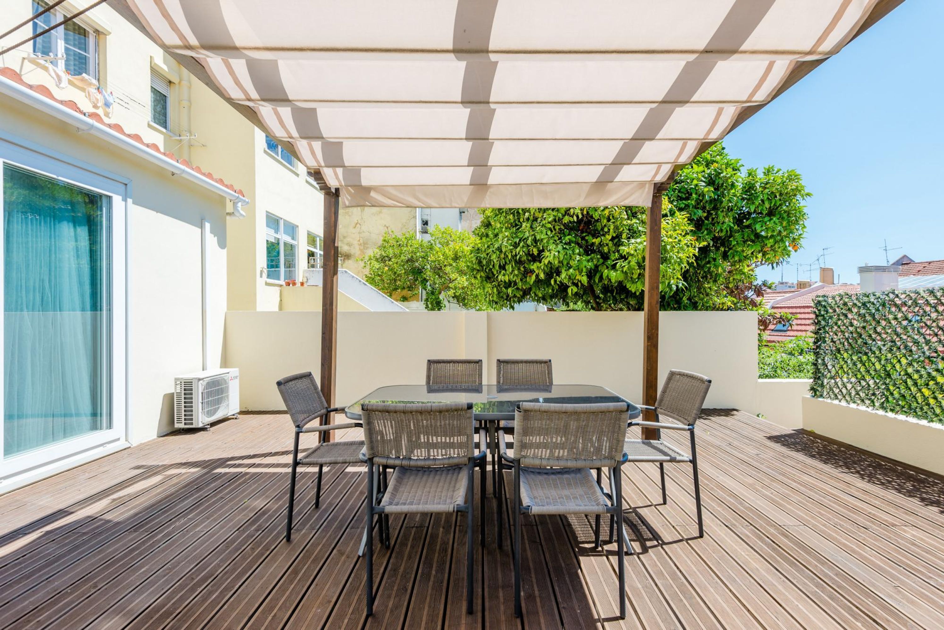 An outdoor patio with a table and chairs, covered by a canopy, and surrounded by greenery.