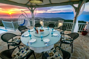 A round table set for four with chairs, a hanging chair, and a view of the sea at sunset.