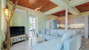 A spacious living area with a large light blue sofa, a flat-screen TV on a wooden stand, and a dining area with wooden cabinets and a kitchen counter. - Villa Orizontas Corfu, Private Villa With Breathtaking Views (Corfu)