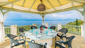 An outdoor dining area with a table set for four, surrounded by wicker chairs, overlooking a scenic view of a lake and mountains. - Villa Orizontas Corfu, Private Villa With Breathtaking Views (Corfu)
