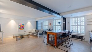 Un vaste espace de vie comprenant une table à manger, un canapé et des fauteuils, de grandes fenêtres équipées de stores, et une cuisine moderne en arrière-plan.