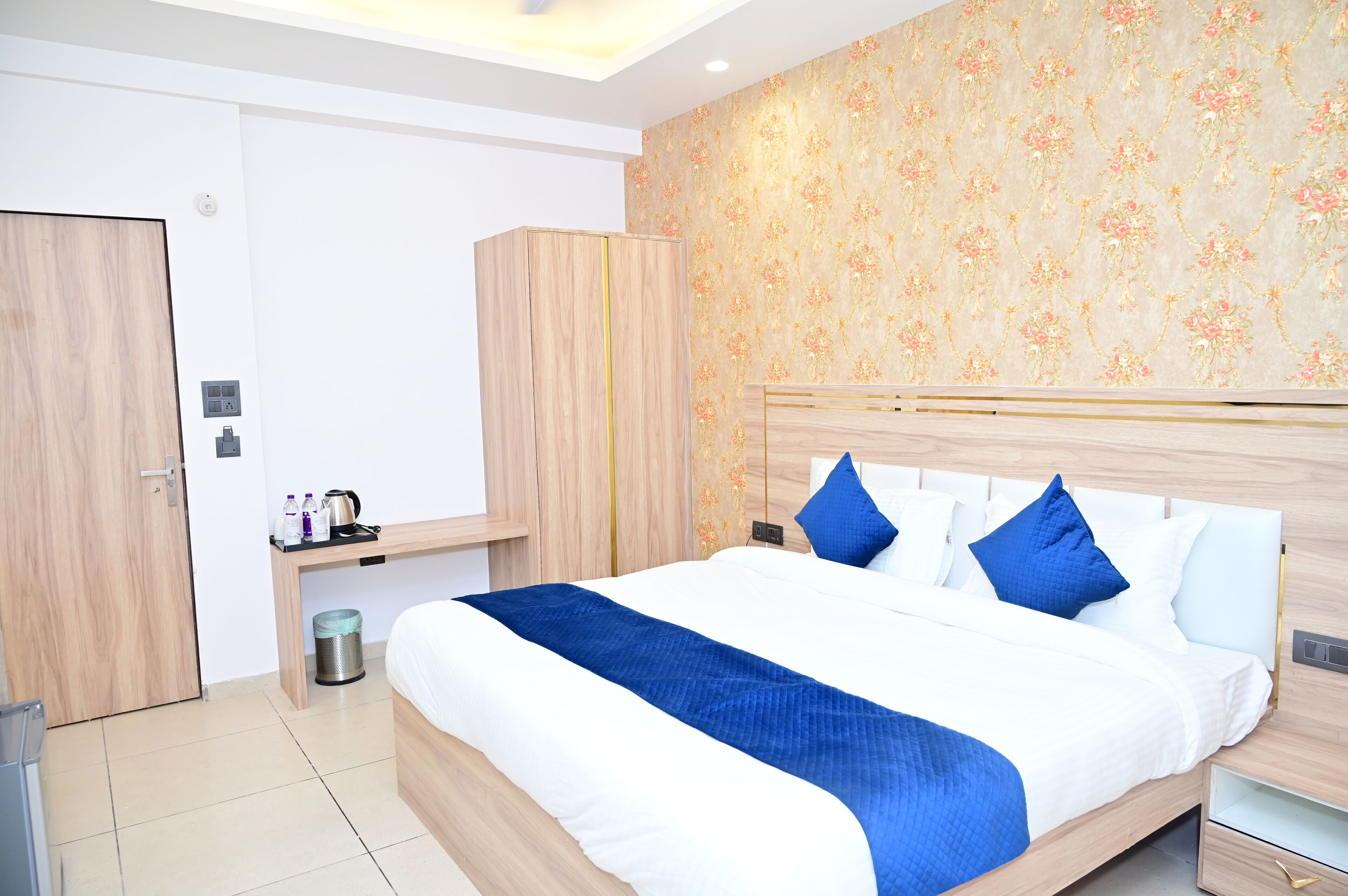 A hotel room with a large bed, a wooden wardrobe, a desk with a chair, and a small trash bin.