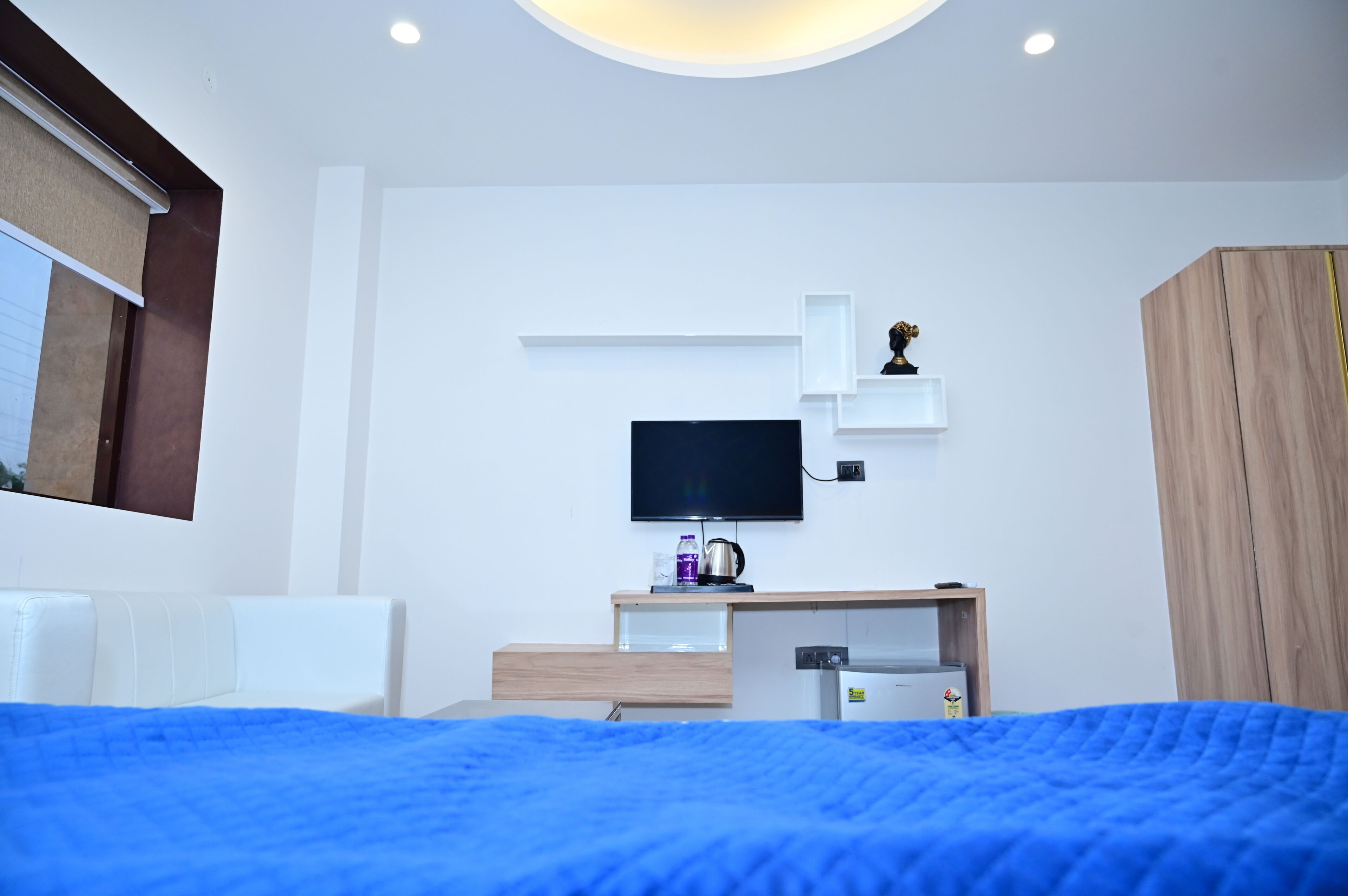 A modern hotel room with a bed, a TV, a small desk with a kettle, and a wardrobe.